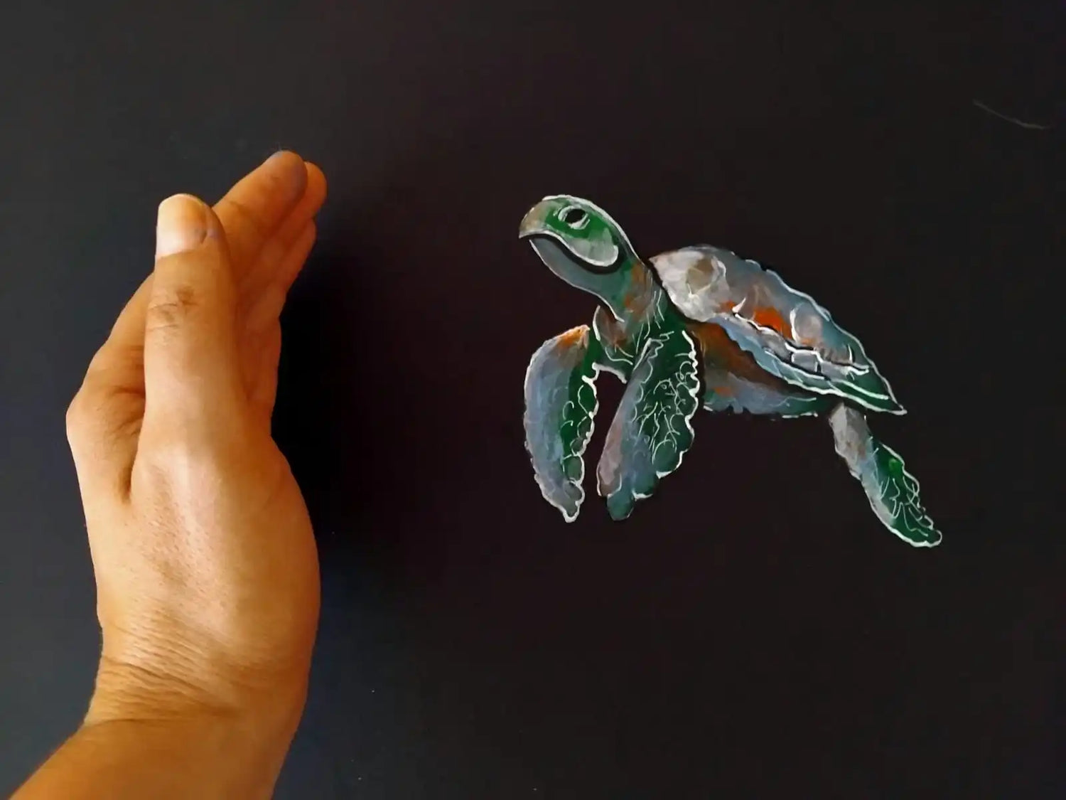 A hand next to a painted sea turtle on a dark surface.